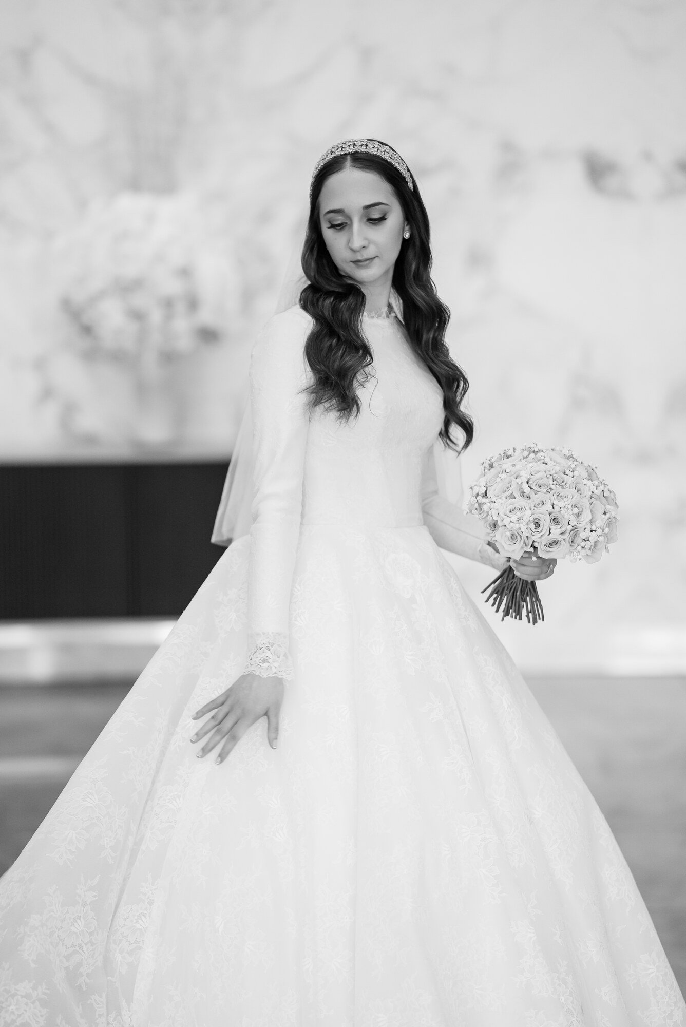 Black and white full-length portrait of the bride in a long-sleeved lace gown holding a pale rose bouquet against a marble wall