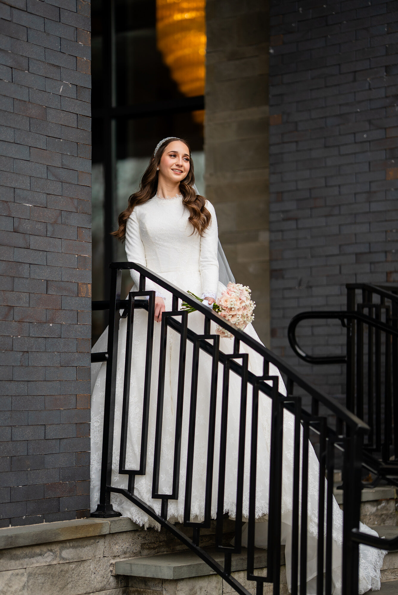 The bride in a long-sleeved lace gown and crystal headband on a staircase, holding a blush rose bouquet, grey brick behind