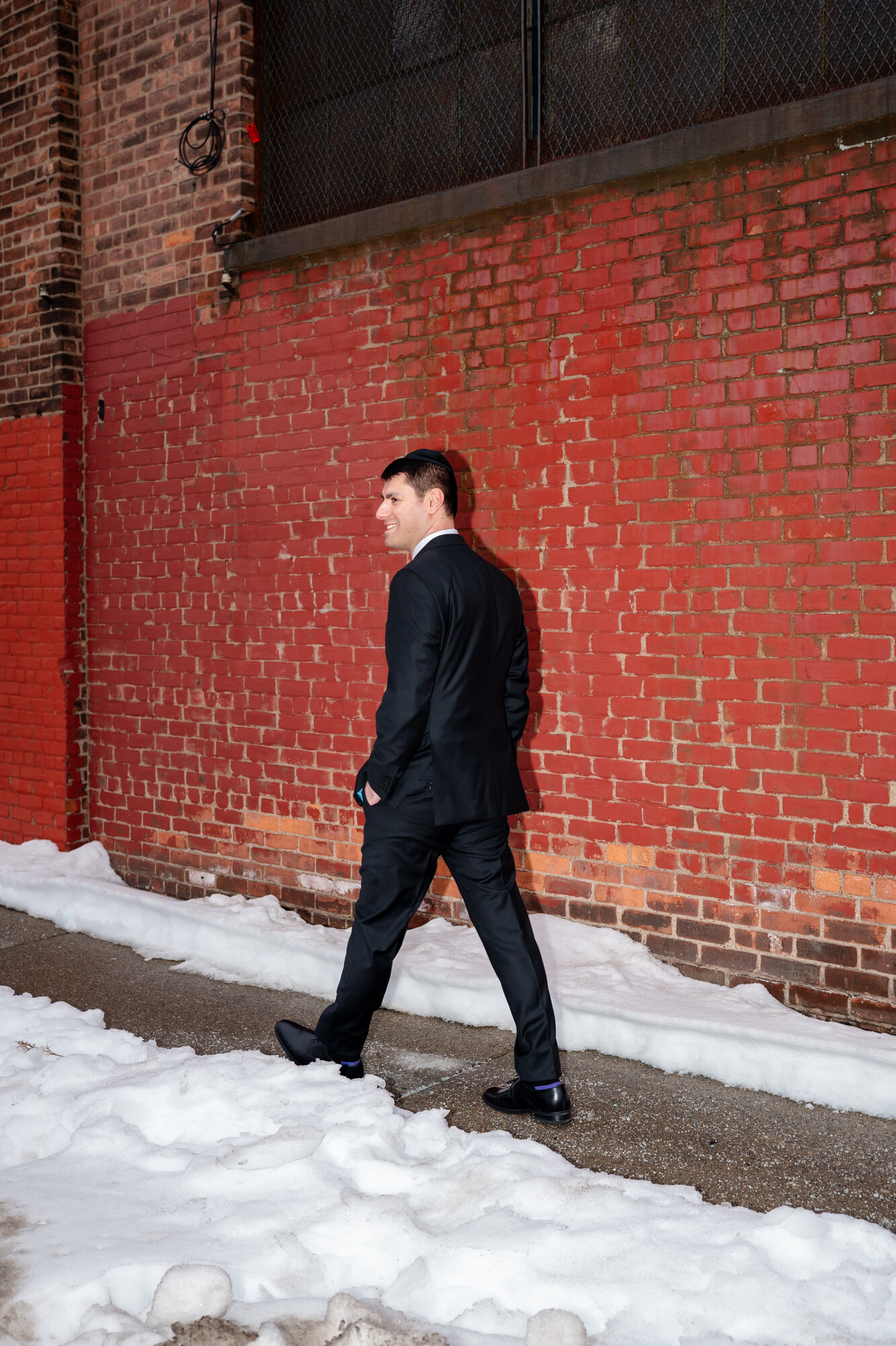 The groom in a dark suit and kippah glancing back over his shoulder as he walks past a red brick wall with snow on the ground