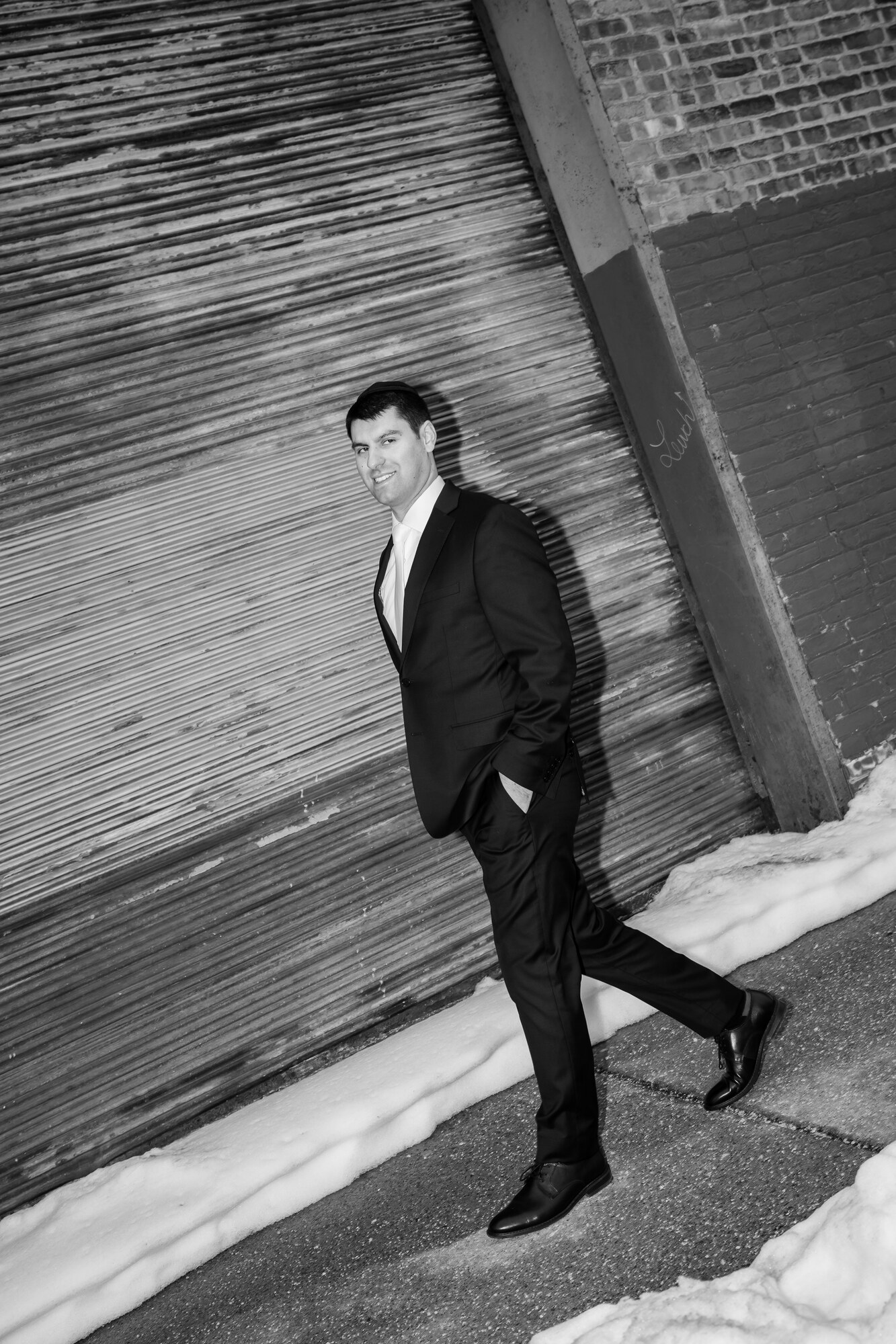 Black and white shot of the groom walking past a metal roll-down gate, snow along the kerb, looking toward the camera