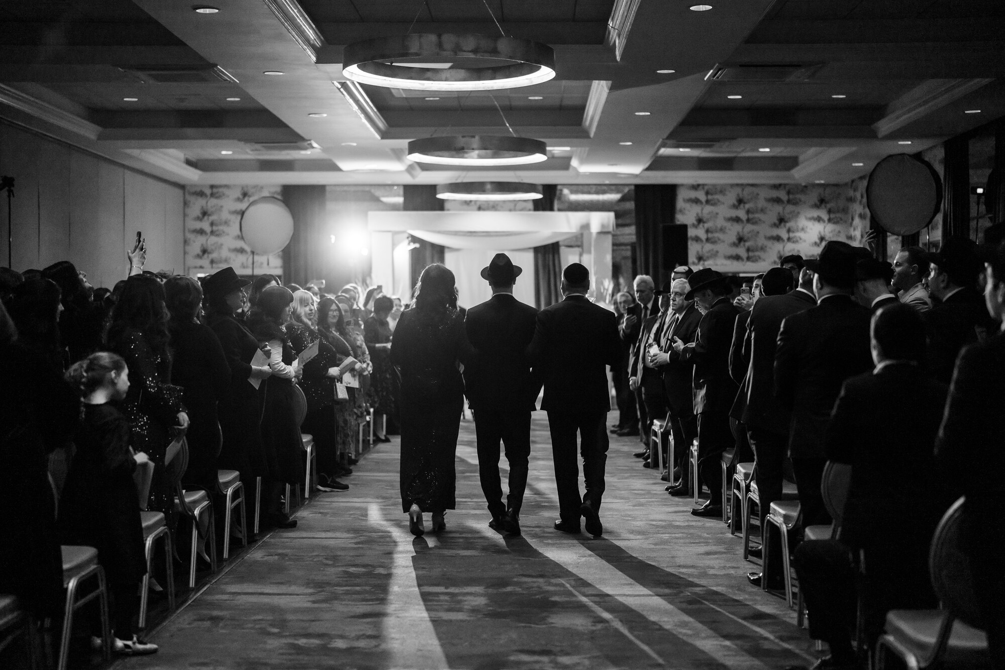 Black and white view down the aisle as the groom walks in between his parents, guests standing on both sides toward the lit chuppah