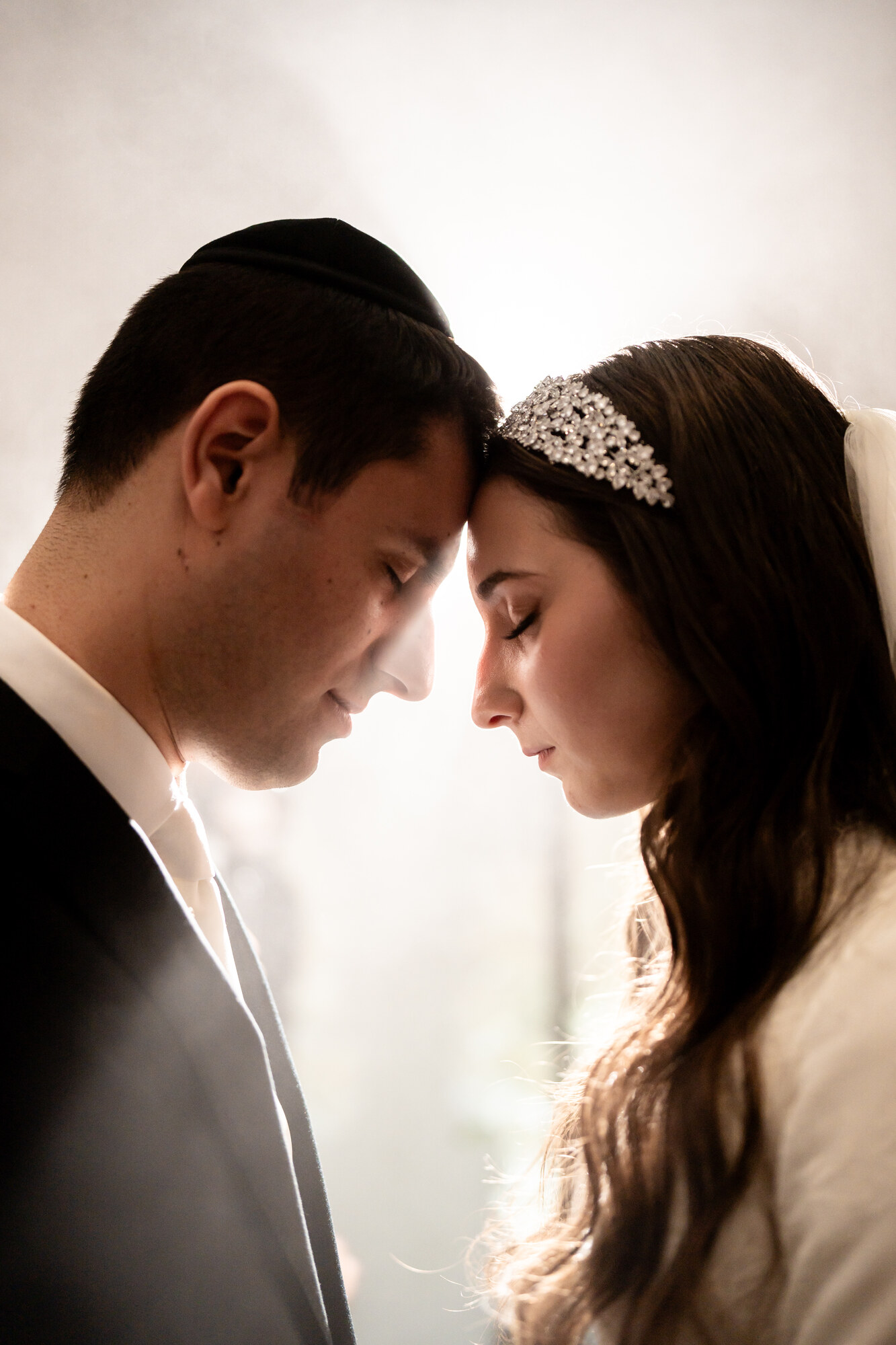 Backlit black and white portrait of the bride and groom forehead to forehead with their eyes closed, window light behind them