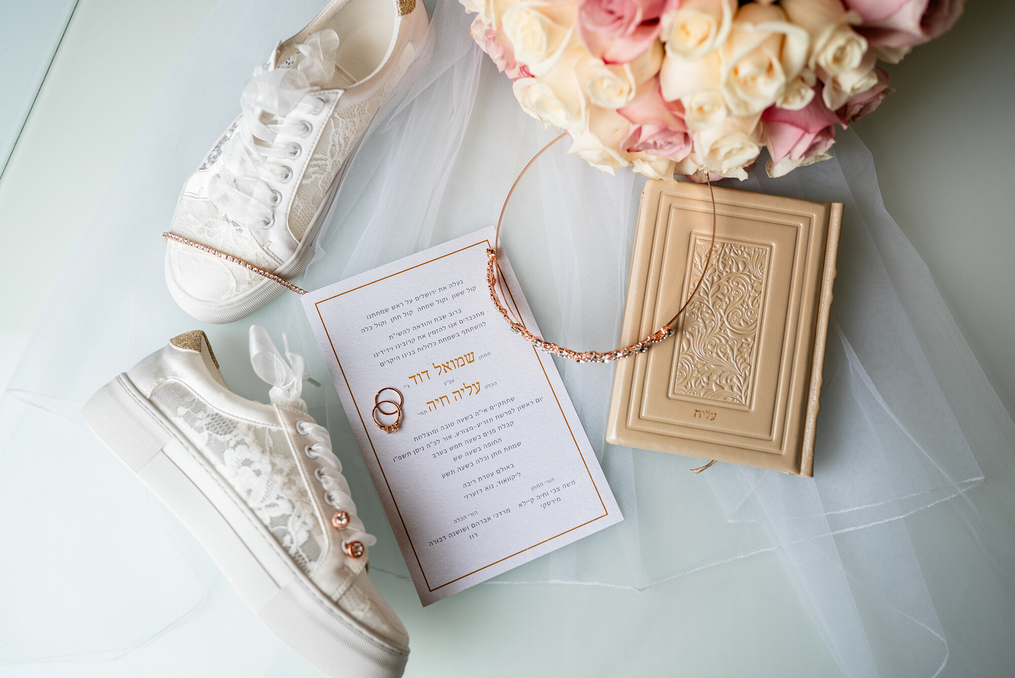 Flat lay on pale blue of white lace platform sneakers, a Hebrew invitation with two rings, a crystal headband, a leather bencher and blush roses