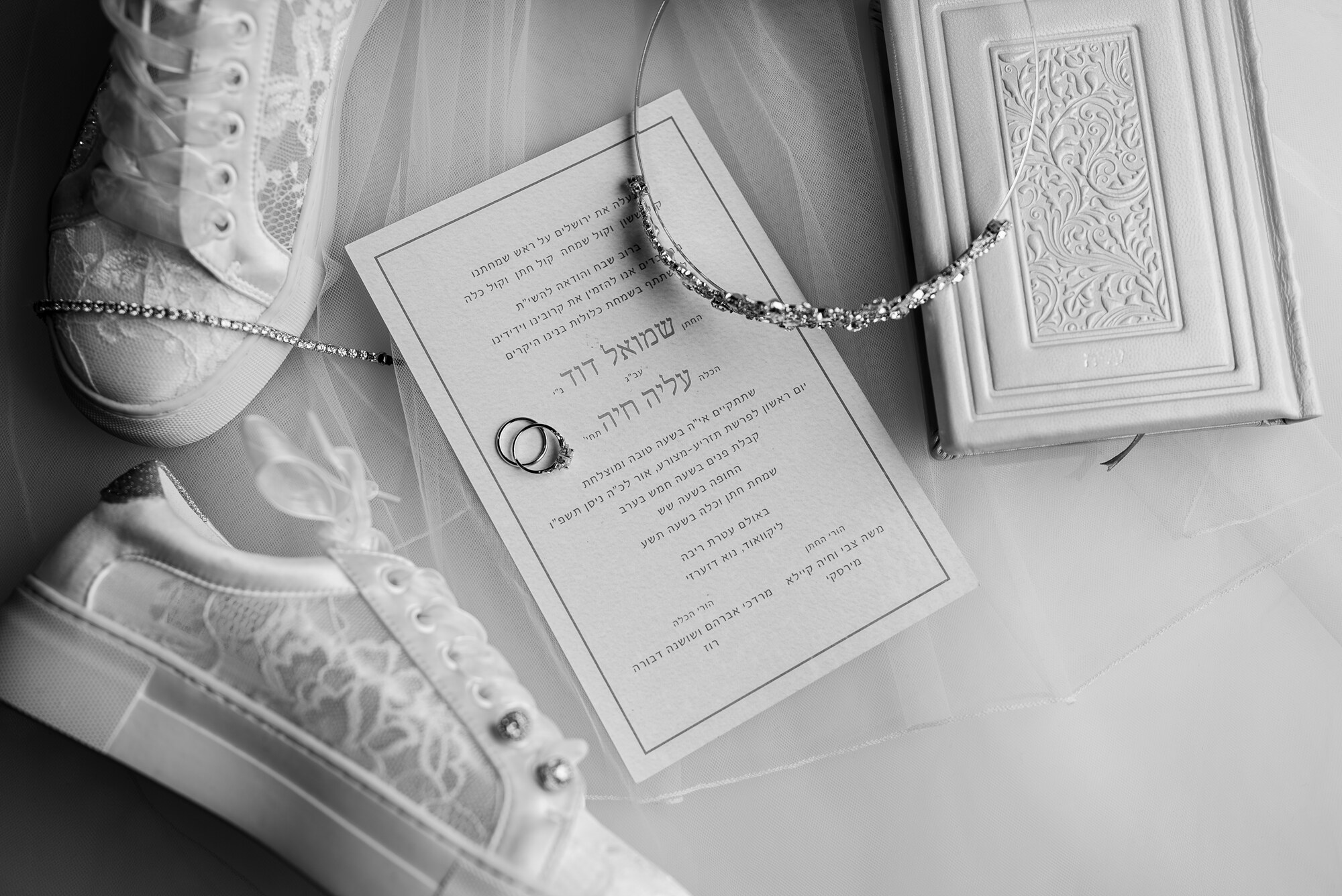 Black and white flat lay of lace bridal sneakers, a Hebrew invitation with rings, a crystal headband and an embossed bencher on tulle