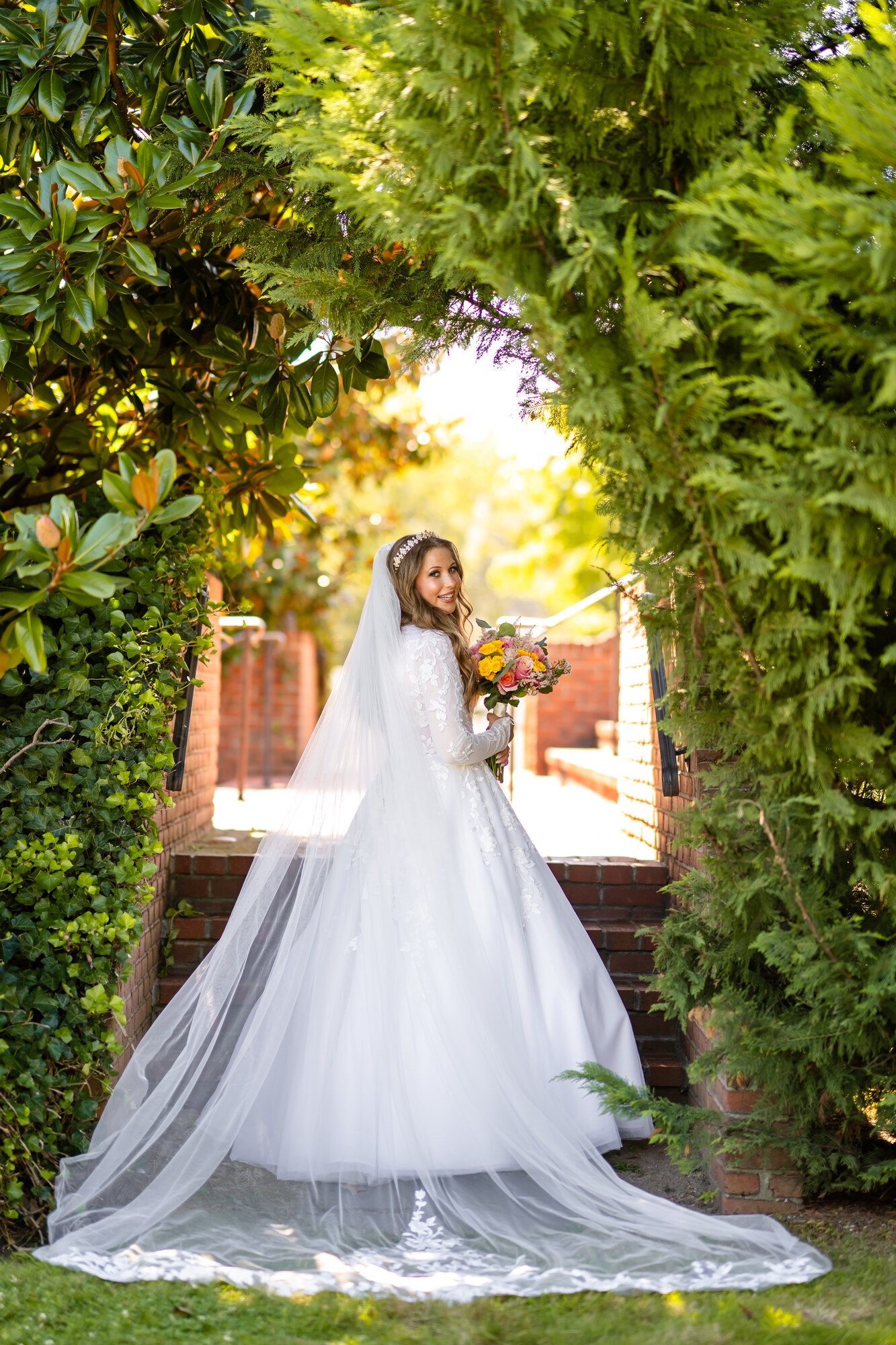 The bride standing beneath an arch of magnolia and evergreens at the foot of brick garden steps, her long lace-edged veil and train spread across the lawn, sun breaking through behind