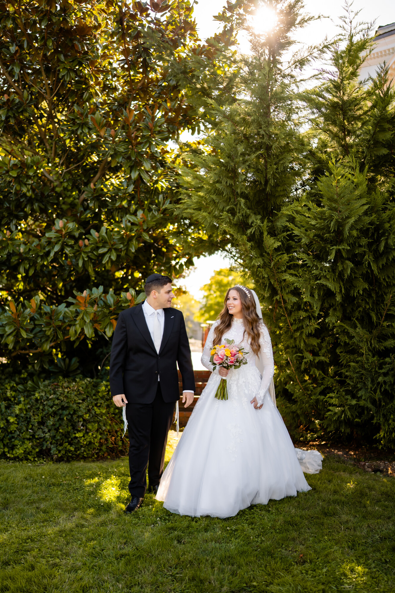 The bride and groom walking toward the camera on a sunlit garden path lined with tall evergreens, strong sun flare overhead