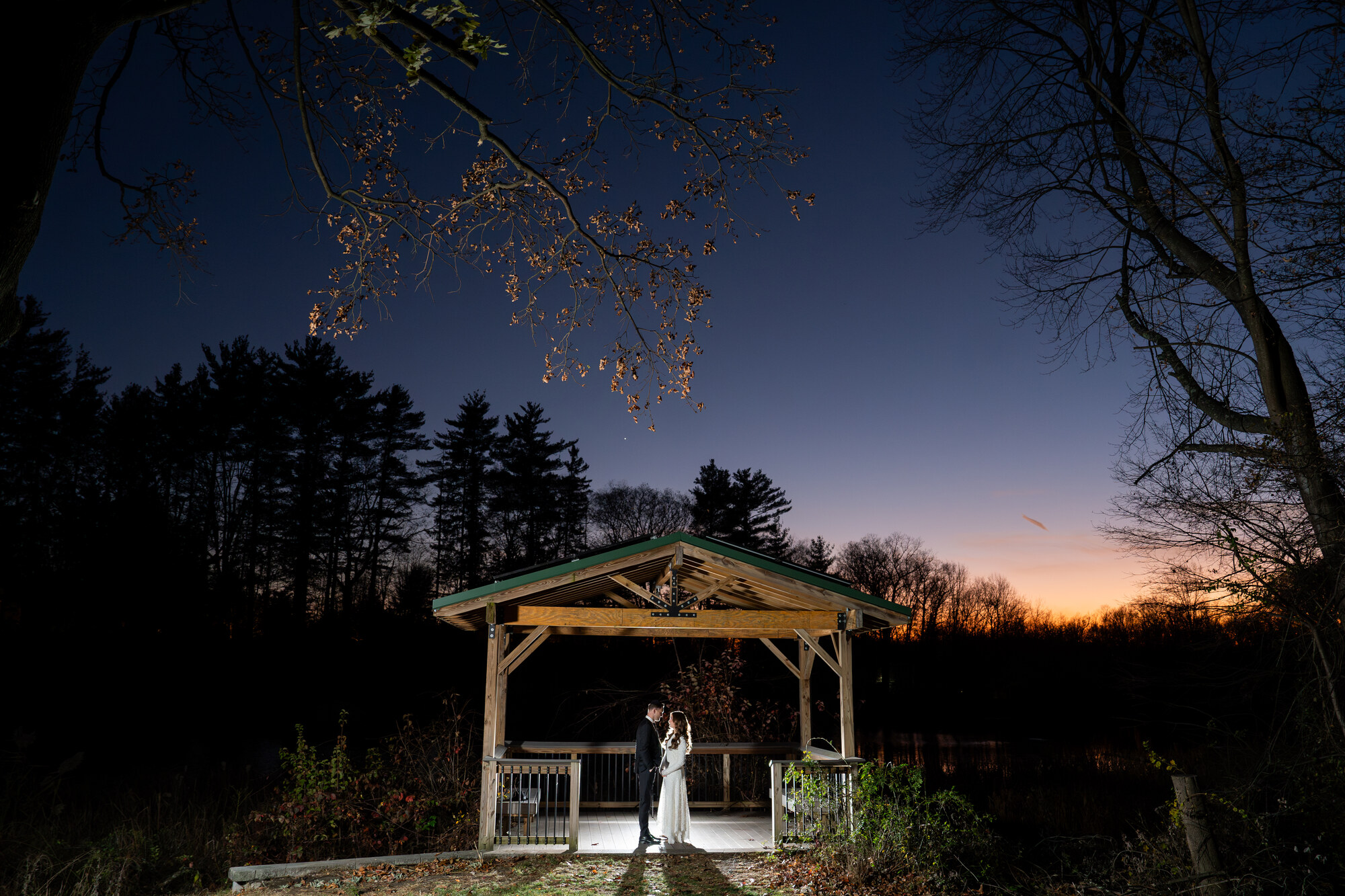 Blue-hour portrait of the bride and groom facing each other inside a lit wooden gazebo by a pond, orange sunset glow on the horizon