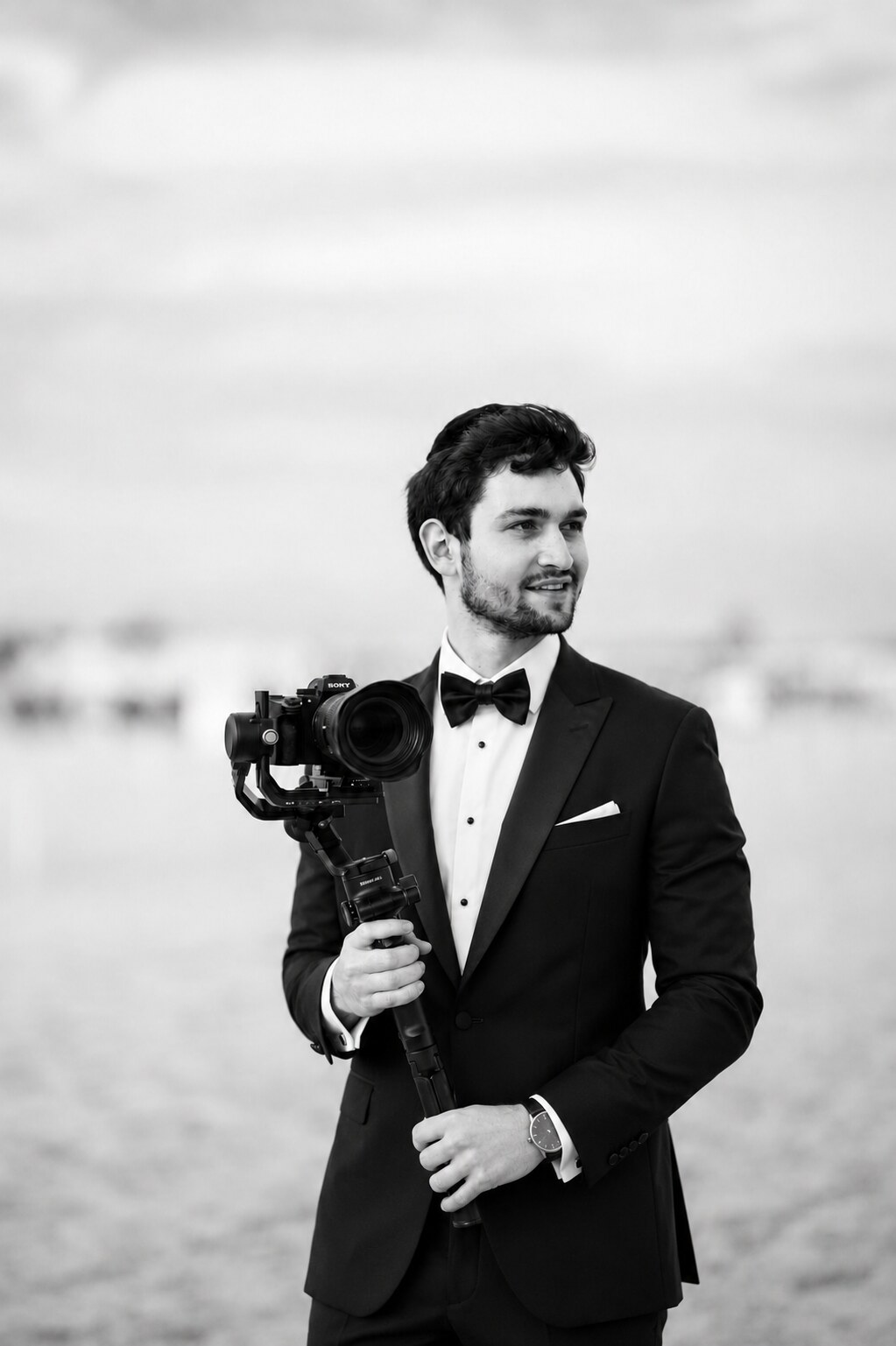 Black and white portrait of Yaacov King in a tuxedo and bow tie holding a camera on a gimbal, soft out-of-focus background