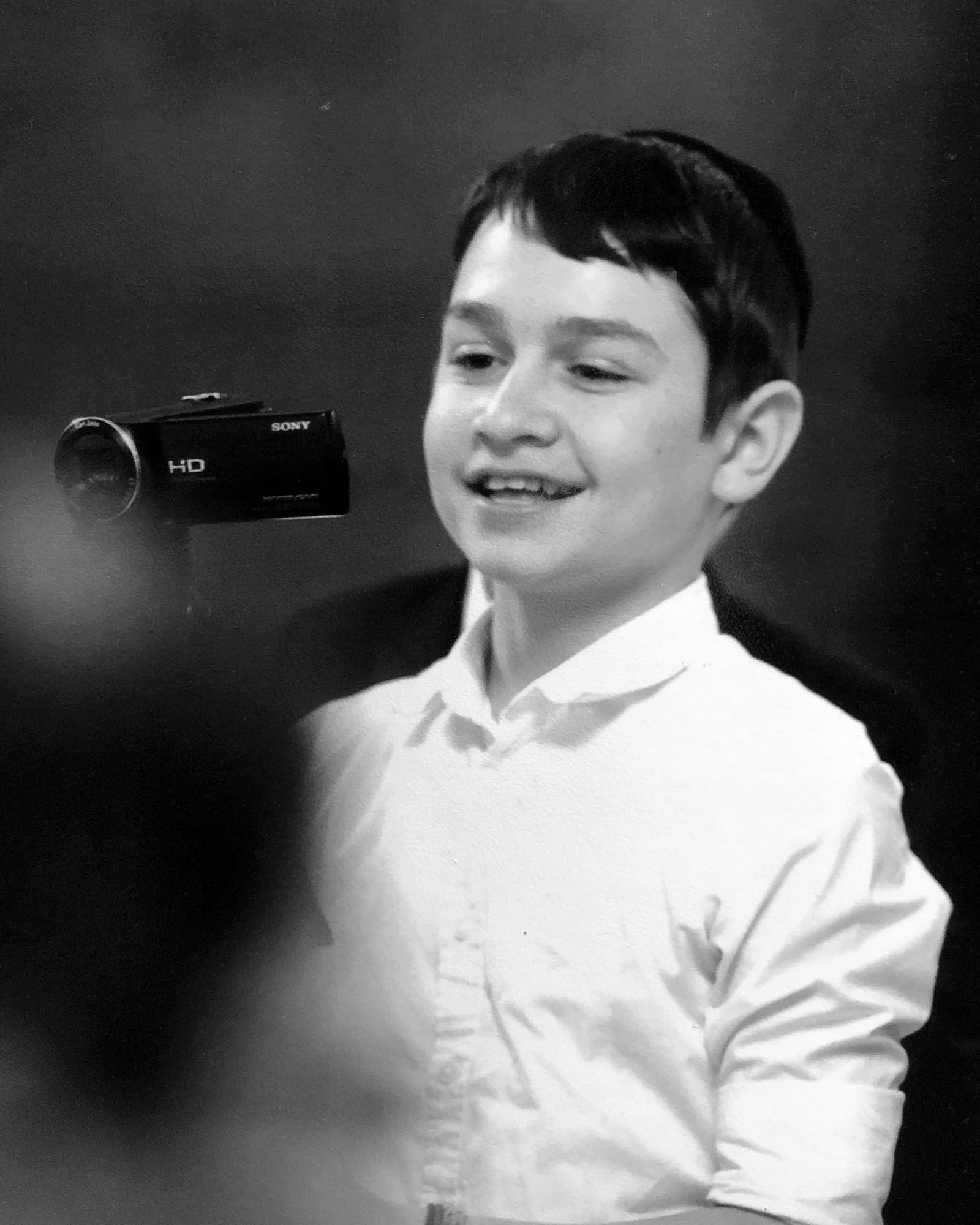 Black and white photo of Yaacov King as a boy in a white shirt, smiling beside a Sony camcorder on a stand